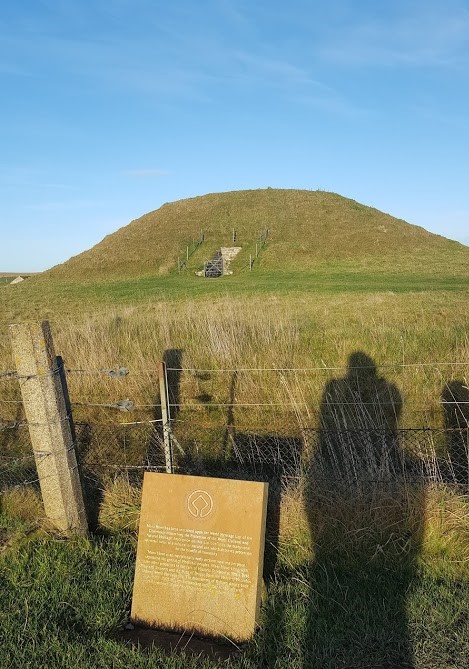 grass field with Maeshowe mound in focus in front is a Heart of Neolithic Orkney info sign