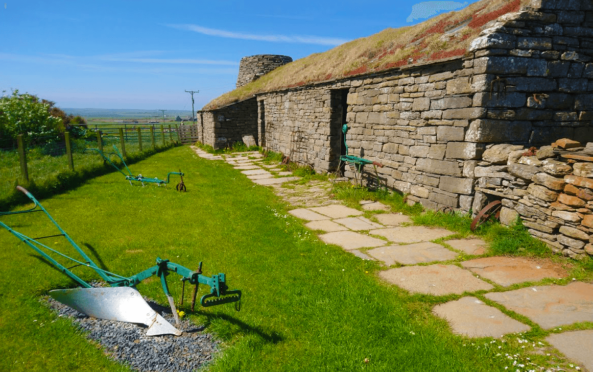 sunny day at the corrigall farm museum ploughs lie on the grass next to stone farmhouse